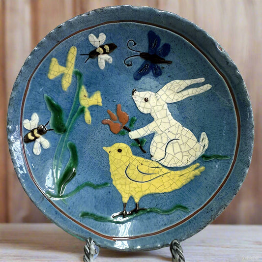 A 6-inch round ceramic plate with a blue background featuring a colorful chick, butterfly, rabbit, and flowers design.
