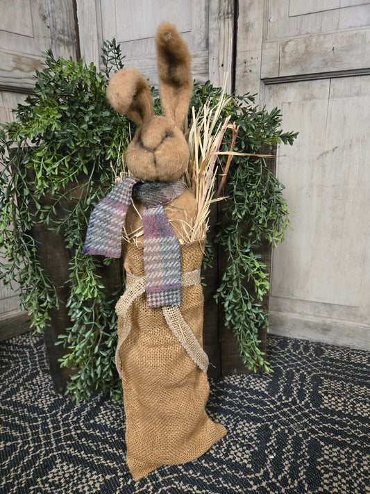 Decorative bunny figure with a plaid bow tie and burlap over a green wreath on a textured surface.