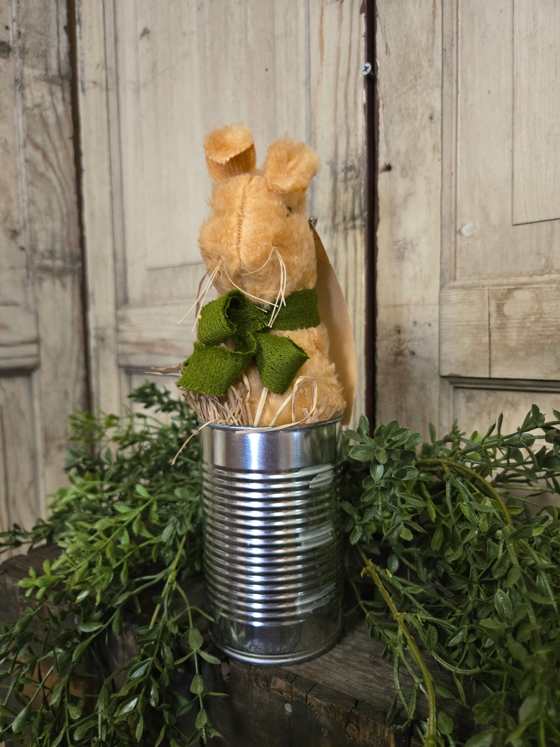 Decorative arrangement with a metal can, greenery, and a small teddy bear on a wooden surface.