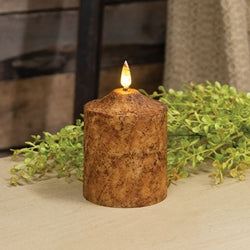An electric pillar candle with a rustic wax coating in a warm burnt mustard shade, featuring a realistic flame that flickers. The candle is placed on a table with a green plant beside it.