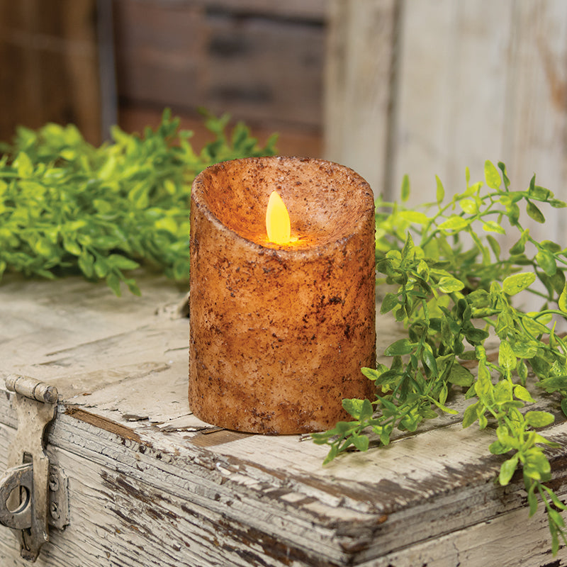 An electric pillar candle with a rustic wax coating in burnt ivory color, featuring a realistic flame, placed on a wooden surface with greenery in the background.