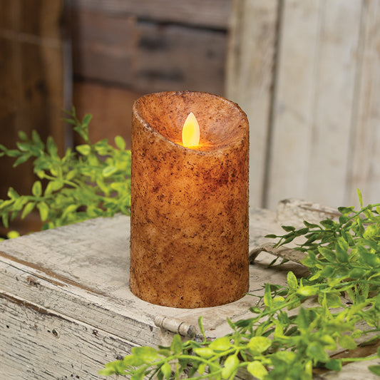 An electric pillar candle with a rustic wax coating in burnt ivory, featuring a realistic flame that is lit, placed on a wooden surface with greenery in the background.