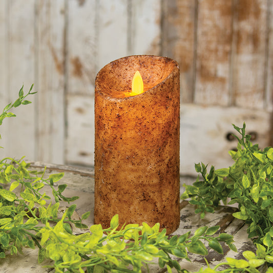 A battery operated pillar candle with a rustic wax coating in a warm burnt ivory shade, featuring a realistic flame that is lit, placed on a wooden surface with greenery in the background.