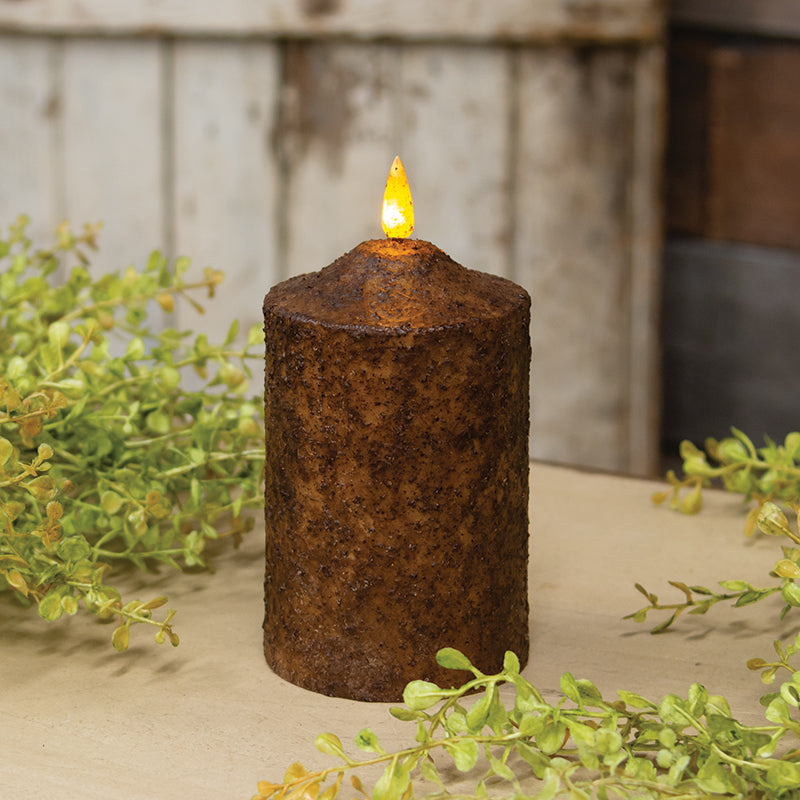 Rust-colored candle with a lit wick on a wooden surface with greenery