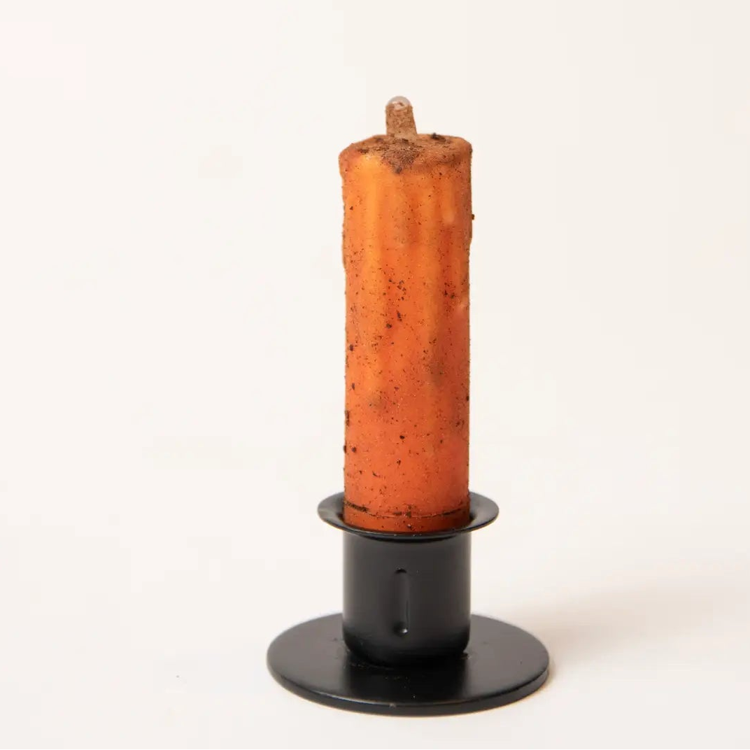 An orange taper candle with a rustic, textured surface sits in a matte black metal candlestick holder against a clean, off-white background.