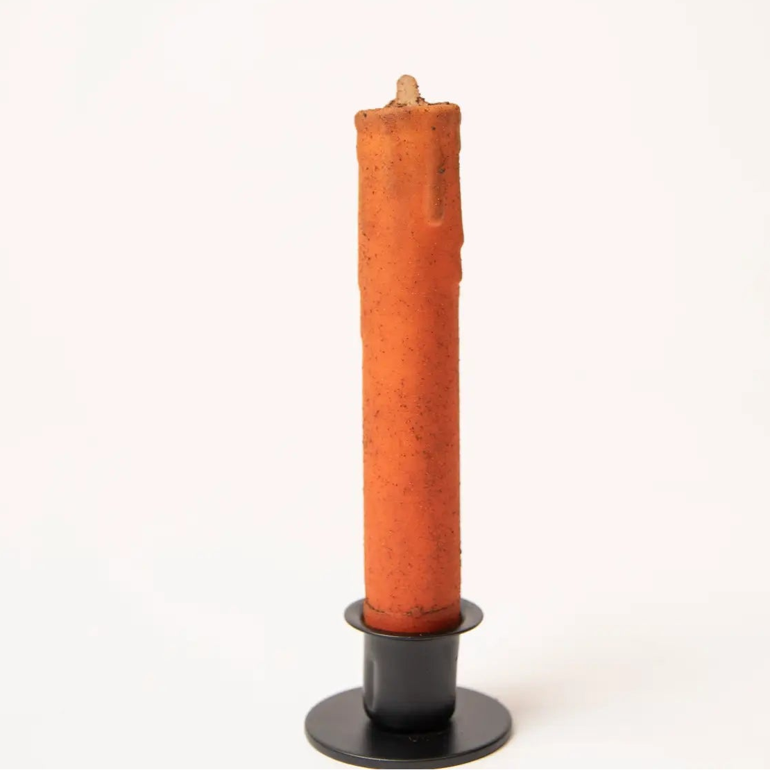 An orange taper candle with a textured, melted wax finish, standing in a black metal holder against a plain white background.