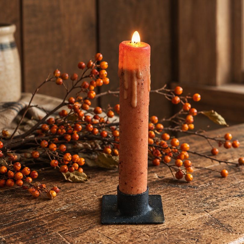 A lit orange taper candle with a black metal base sits on a rustic wooden table, surrounded by orange berries and a cozy wood-paneled background.