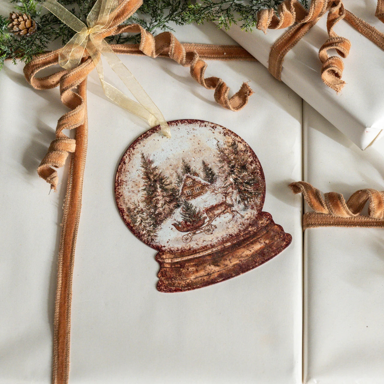 A round vintage-style snow globe holiday ornament with a snowy winter scene, displayed on white wrapped gift boxes with tan ribbon and greenery.