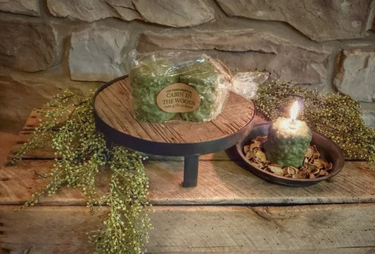 Two greenery-infused votive candles on a wooden platter with a label reading 'Cabin in the Woods', placed on a wooden surface with decorative stones and a candle lid nearby.