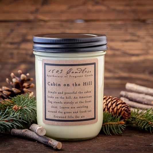 A beige jar candle labeled 'Cabin on the Hill' with a capacity of 14 ounces, displayed on a wooden surface with pine needles and decorative branches around it.