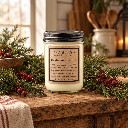 A glass jar candle with a black lid and a cream-colored wax body sits on a rustic wooden table, surrounded by pine branches, red berries, and a striped cloth. The label reads '1965 Farmhouse Apothecary of Fragrant Goods' and 'Cabin on the Hill'.