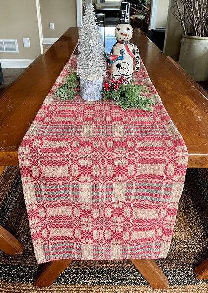 A rectangular woven table runner with a red, beige, and blue Nordic-style geometric pattern, styled on a wooden dining table with holiday decorations.