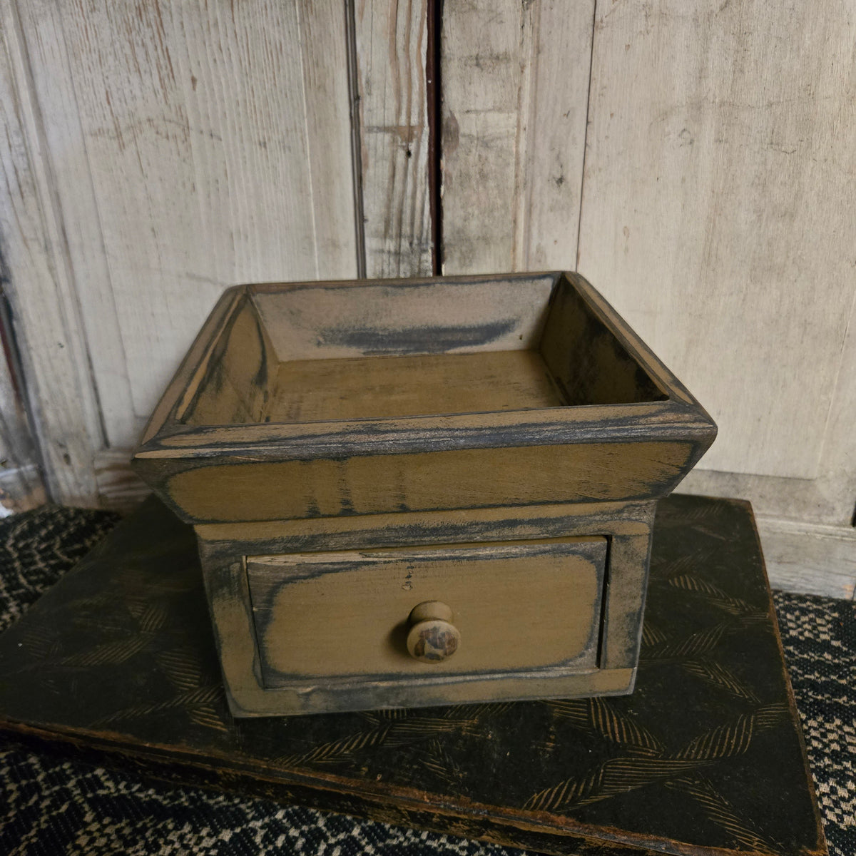 A rustic, distressed wooden drawer box with a single round knob, set against a weathered white wooden wall background.
