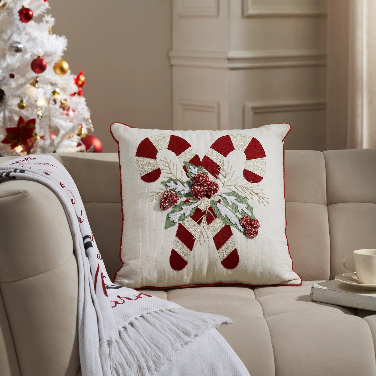 Candy cane and holly Christmas pillow on a beige sofa beside a fringed throw and decorated white Christmas tree.