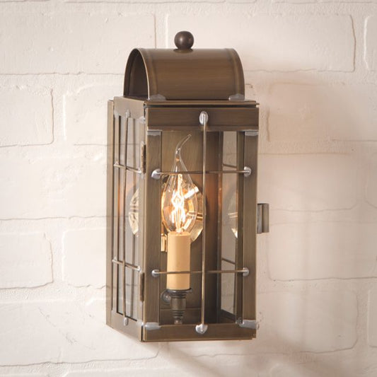 A weathered brass wall lantern with a clear glass front featuring a single light bulb inside, mounted on a white brick wall.