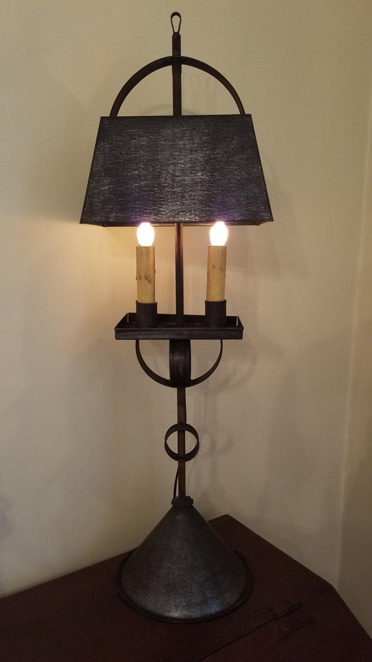 A black tin table lamp with a cylindrical shade, showing two light bulbs, placed on a wooden surface.