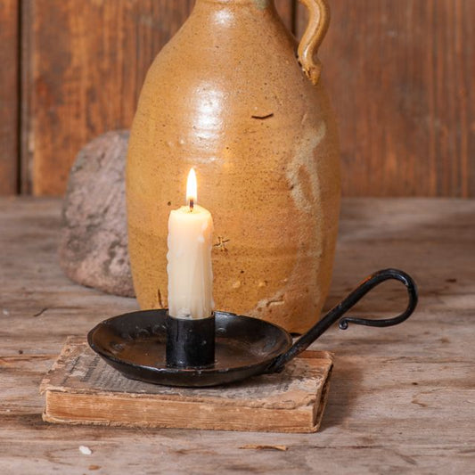 A black cast iron taper candle holder with handles placed on a wooden surface with a lit candle inside it, in front of a beige pottery item.