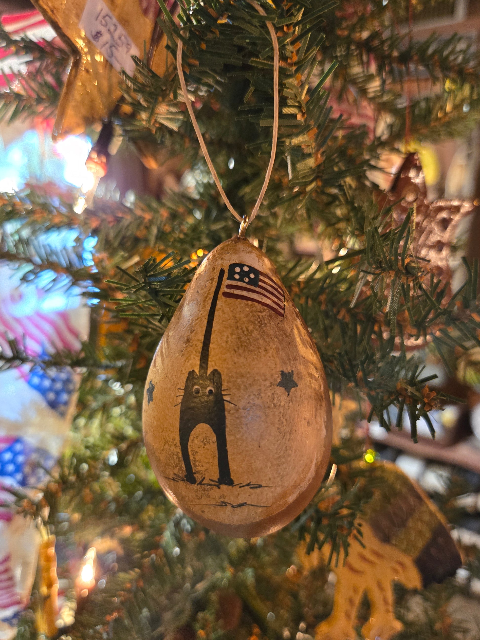 A handcrafted gourd ornament in the painted with a cat and a flag, hanging from a Christmas tree.