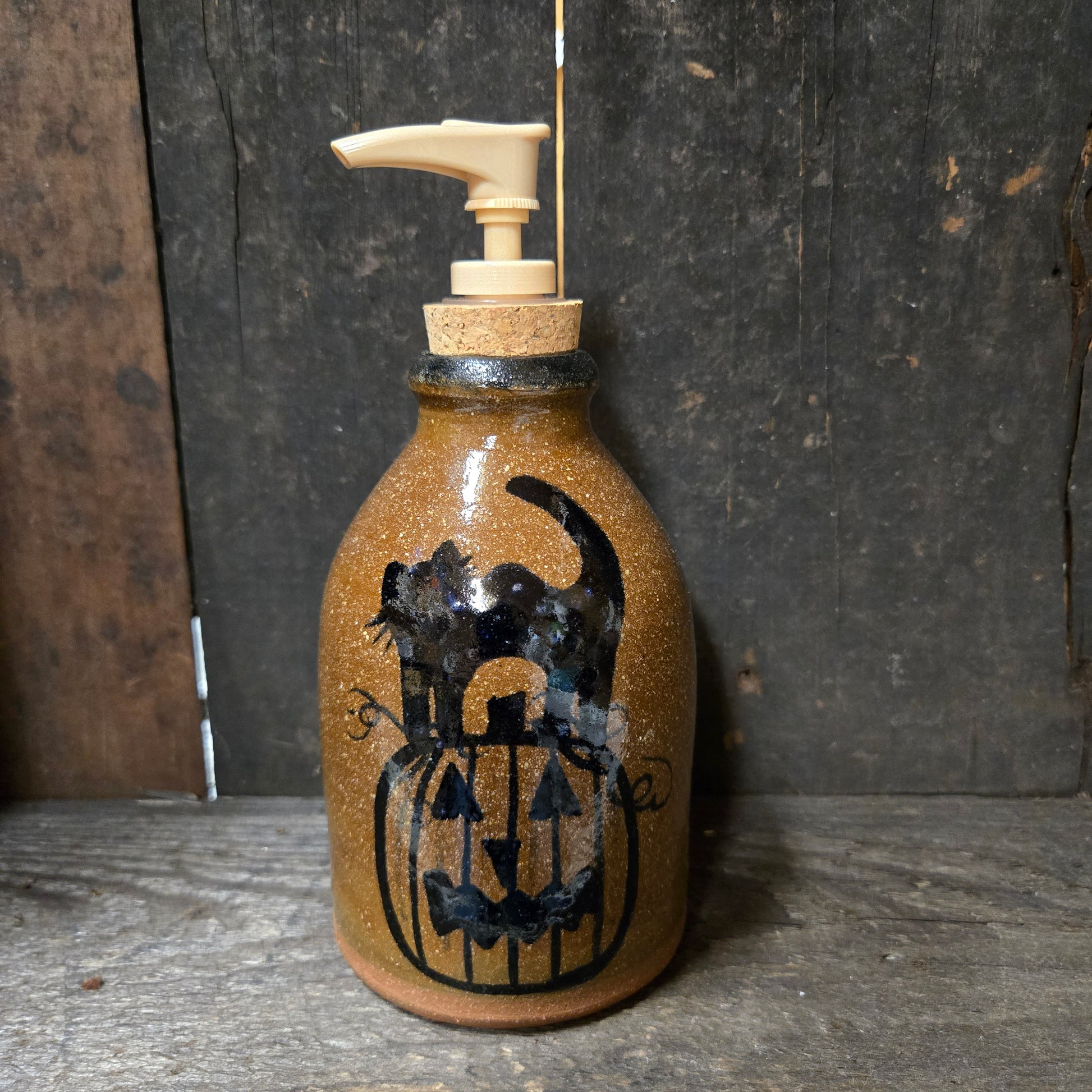 A rustic brown ceramic pump bottle with a black hand-painted jack-o'-lantern and cat design, featuring a cork stopper and cream-colored pump dispenser, set against a dark wooden background.