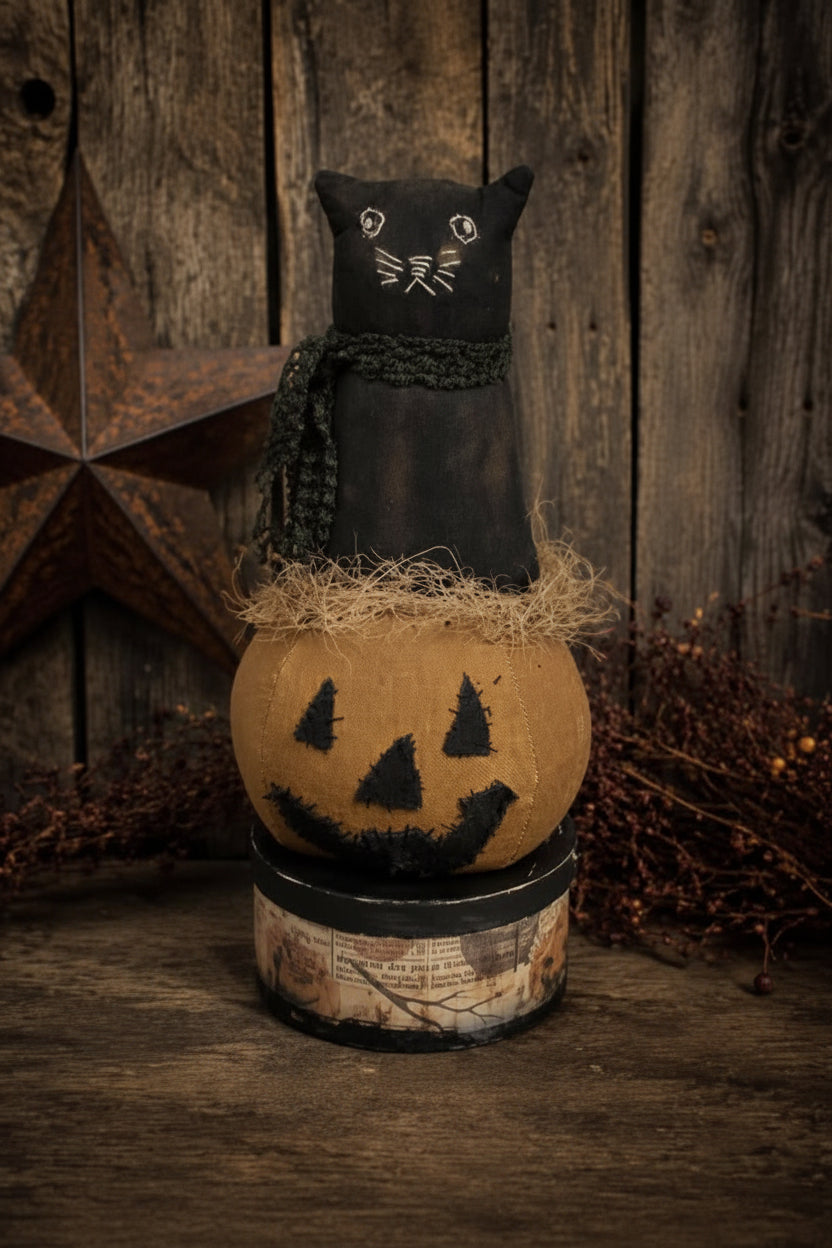 A rustic Halloween decorative piece featuring a black cat figurine sitting on top of a carved jack-o'-lantern pumpkin, displayed on a wooden surface against a weathered wood plank background with a star ornament and dried foliage.