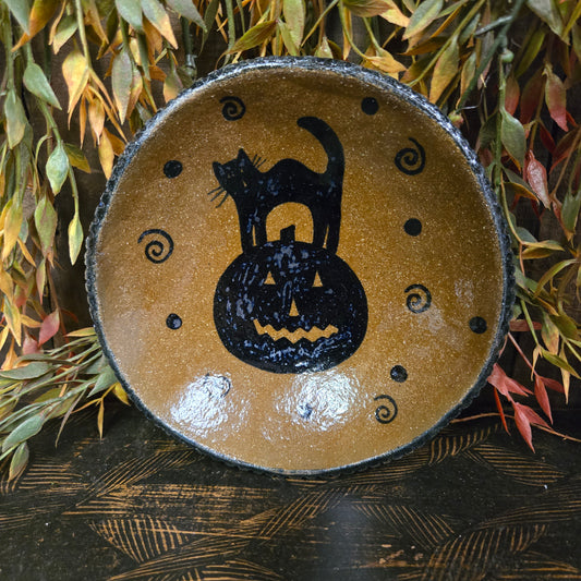 Ceramic plate with black cat and pumpkin design on a wooden surface with leaves.