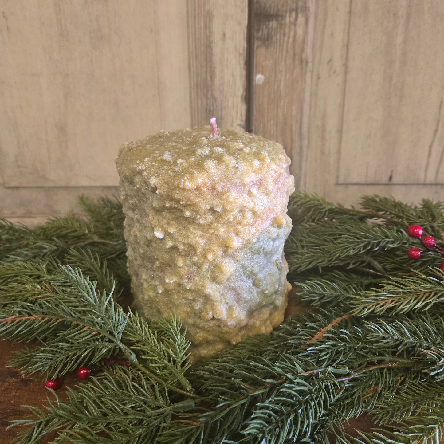 A large hearth candle with a greenish-white wax, possibly cedar and berry scented, placed on a wooden surface with green pine branches and red berries around it.