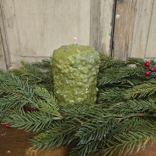 A small pillar candle with a greenish-white wax, placed on a decorative branch, with a wooden background and a small red berry adornment on the side.