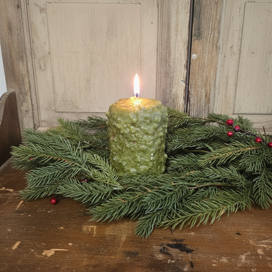 Green textured candle with a flame surrounded by pine branches on a wooden surface.
