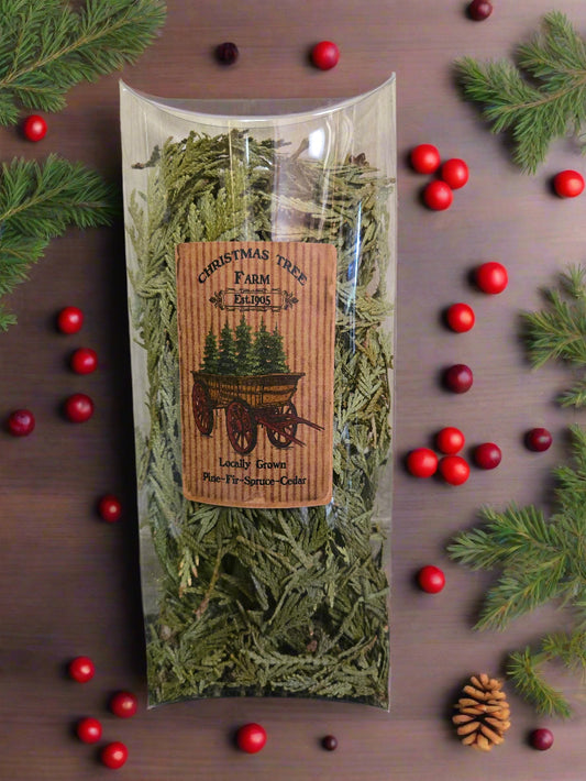 A bundle of cedar tips tied with a string, sitting in a clear plastic container, with a label that reads 'Evergreen Cedar Tips' and 'Primitive Christmas'. The product is placed against a backdrop of a wooden wall and a patterned floor.