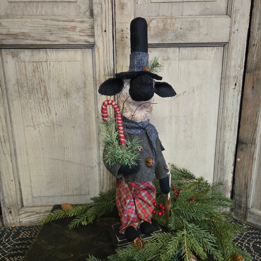Decorative sheep figure with a plaid coat and top hat, surrounded by greenery on a wooden surface.