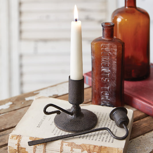 A black cast iron candlestick holder with a taper candle on top, placed on an open book, with a snuffer beside it.