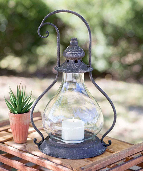 A clear glass candle lantern with a metal frame and a candle inside, placed on a wooden surface with greenery in the background.