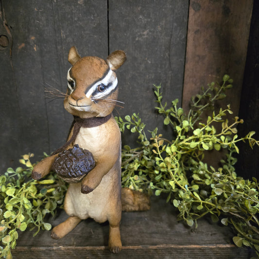 Chipmunk figurine holding a pine cone on a wooden background with greenery