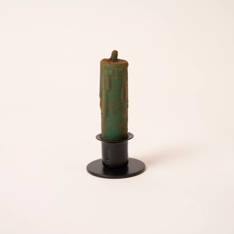 A single candle in a black metal holder on a plain off-white background. The candle has a distressed, weathered green and brown patina finish.