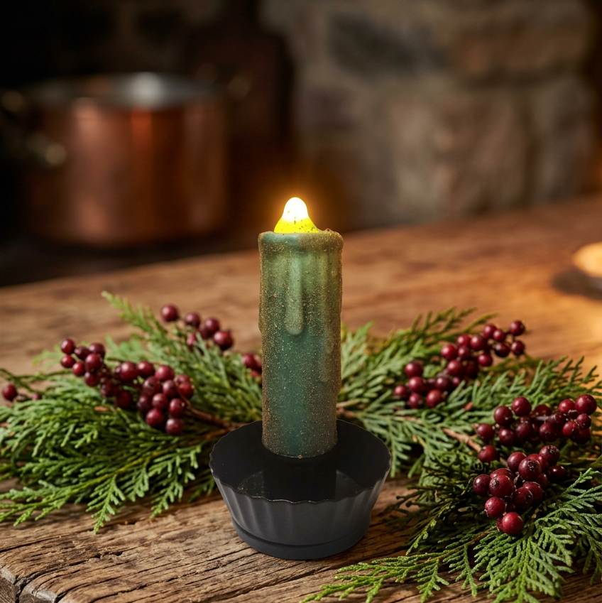 A lit teal-green wax candle in a black holder, styled on a rustic wooden table with evergreen branches and red berries in a cozy indoor setting.