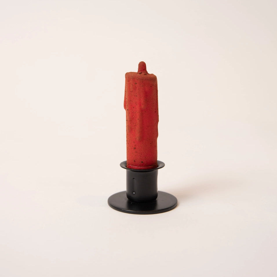 A single red wax candle in a black metal holder, photographed against a plain off-white background.