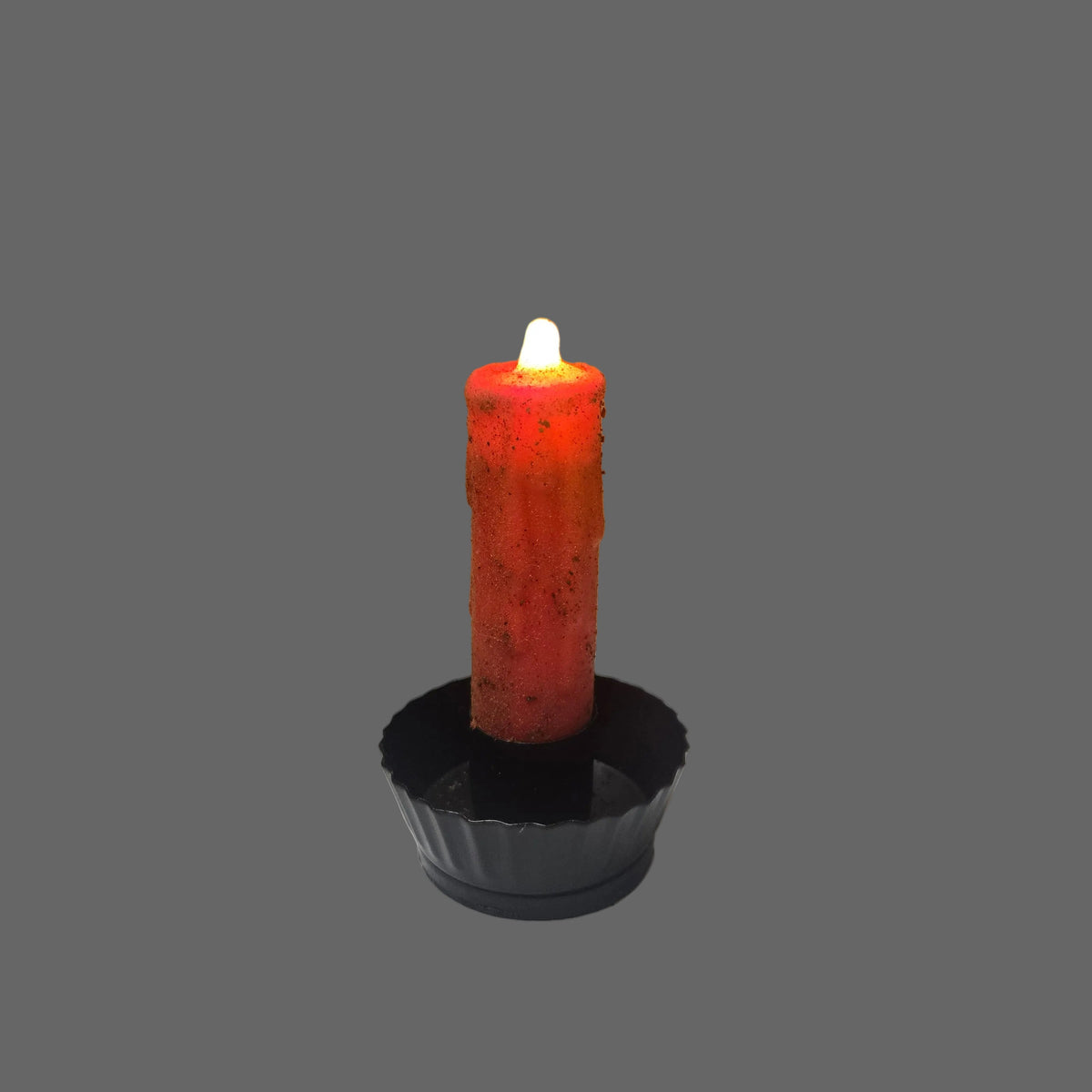 A lit orange-red wax candle in a black plastic cup holder, photographed against a split background of brown corrugated cardboard and a white sheet of paper.