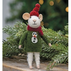Knitted mouse toy wearing a green sweater and red hat with snowman design, standing on a wooden surface with Christmas trees in the background.