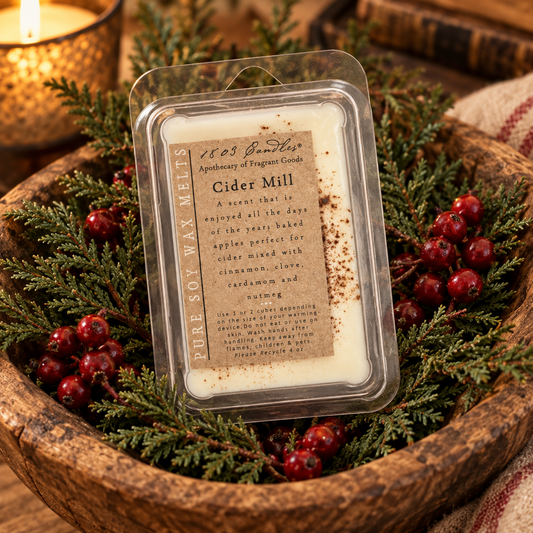 A clear plastic clamshell package of soy wax melts sits in a rustic wooden bowl filled with evergreen branches, red berries, and a lit candle. The label reads '1863 Sweetings Apothecary of Oregon Candles' and 'Cider Mill'.