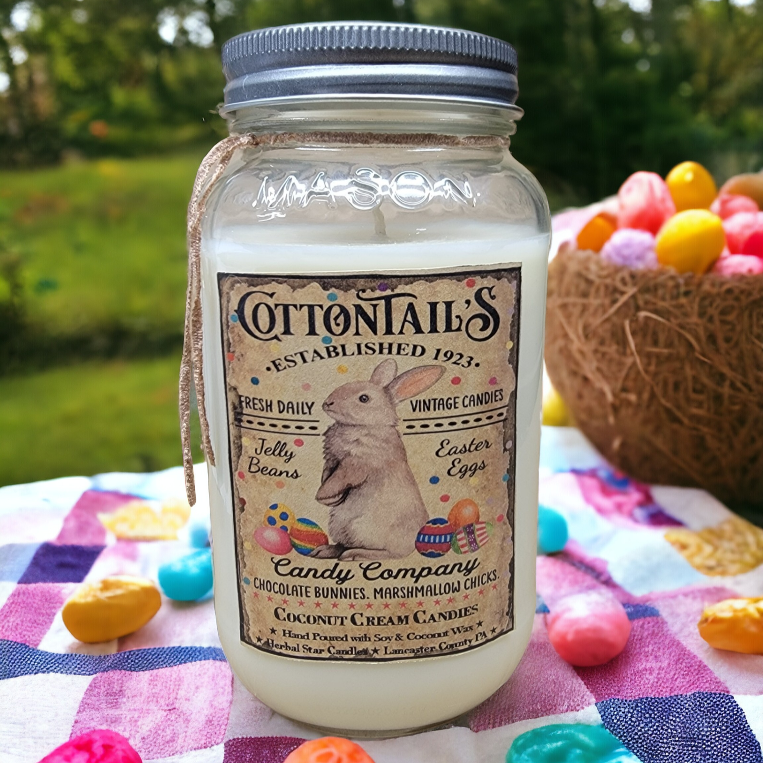 A 24 oz Mason jar candle with a label that reads 'Cottontails Established 1921', featuring an illustration of a rabbit, and listing the scent as Coconut Cream Candies. The jar is placed on a picnic cloth with Easter-themed decorations around it.