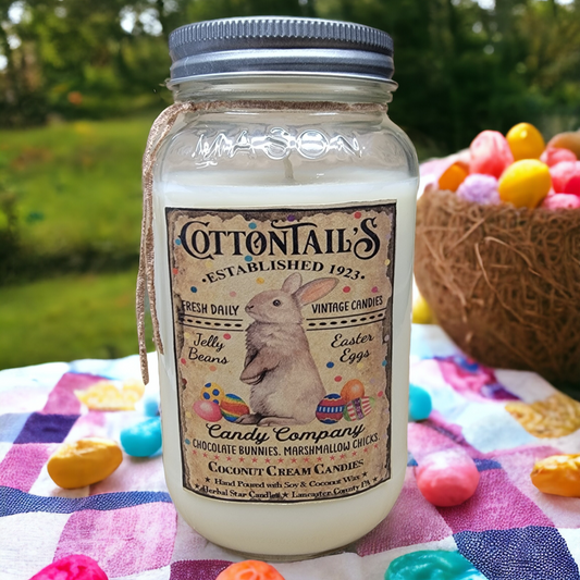 A 24 oz Mason jar candle with a label that reads 'Cottontails Established 1921', featuring an illustration of a rabbit, and listing the scent as Coconut Cream Candies. The jar is placed on a picnic cloth with Easter-themed decorations around it.