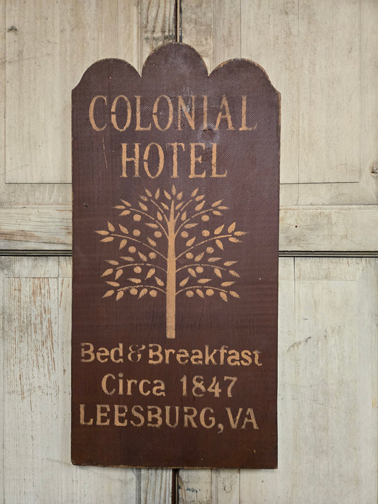 A rustic dark brown wooden sign with scalloped top and engraved text reading 'COLONIAL HOTEL Bed & Breakfast Circa 1847 LEESBURG,VA' plus a tree design, shown against a light weathered wood background.