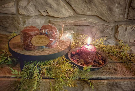 Two candles on a wooden platter with what appears to be a homemade decorative element, placed on a wooden surface with greenery around it.
