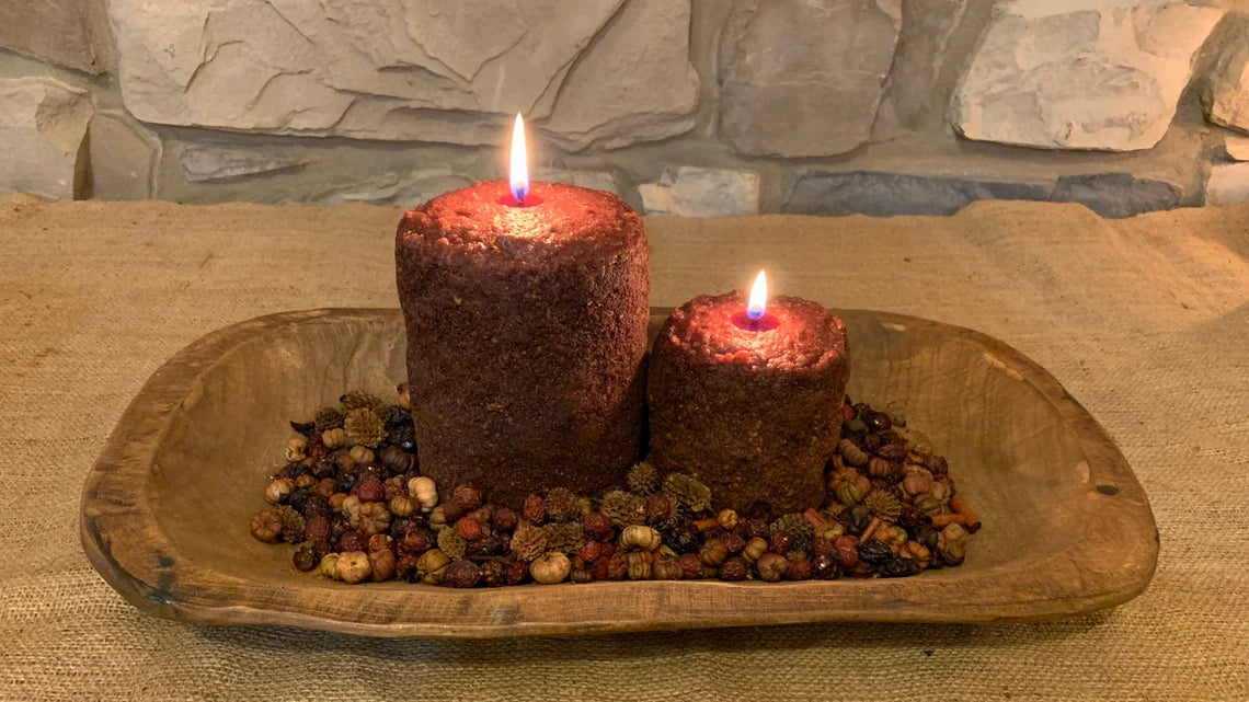 Two cylindrical candles with a rustic appearance, placed on a wooden dish, with a backdrop of a stone wall.