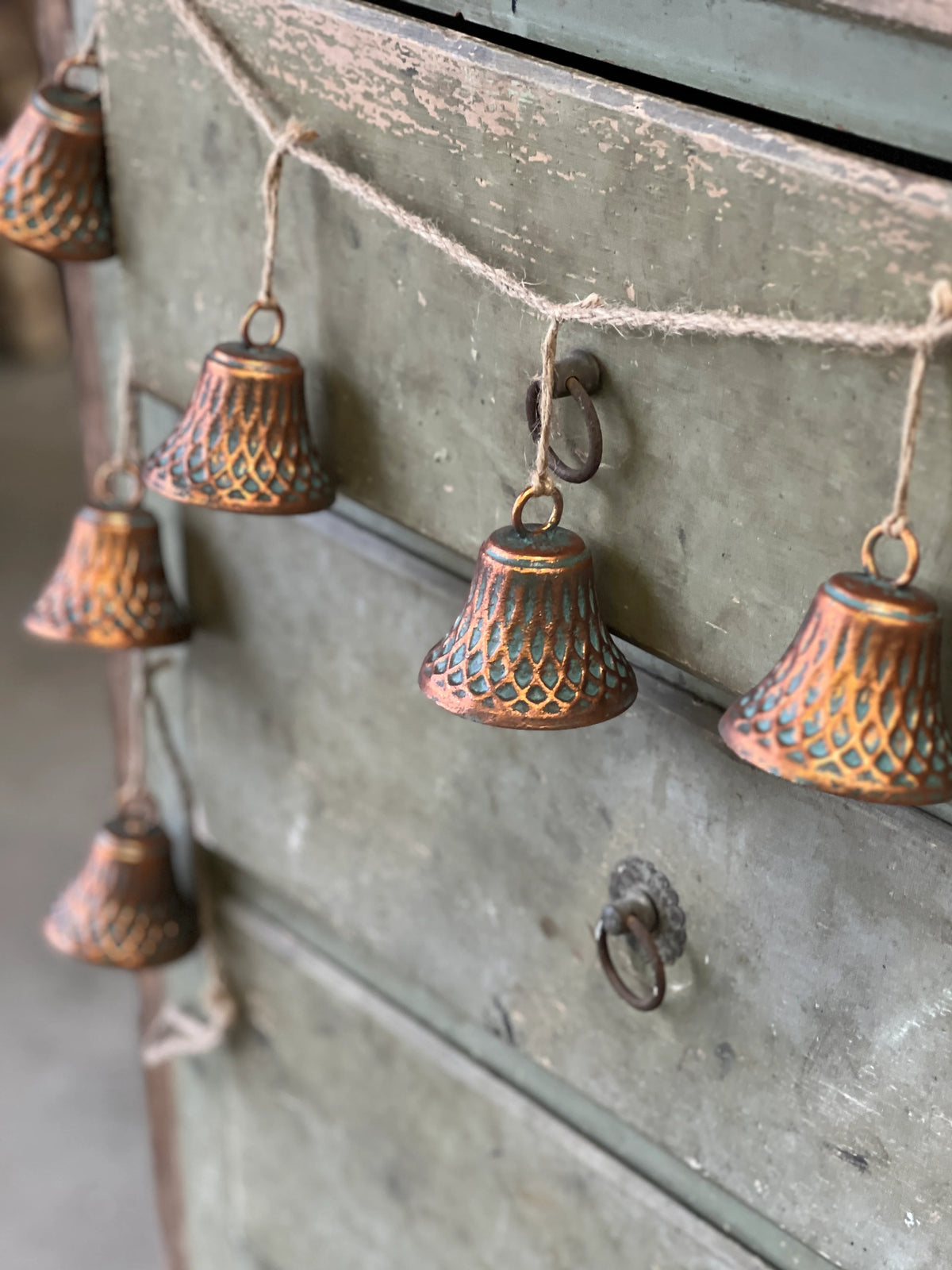 A set of copper-colored bells with a geometric pattern, displayed as a garland and hung over the edge of a green drawer or cabinet.