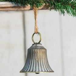 A copper washed liberty bell ornament with a jute rope hanger, lightly whitewashed and with a copper patina finish, displayed against a backdrop of a pine tree and a white wall.