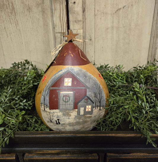 A hand-painted lighted gourd with a country barn scene, featuring an orange roof, white walls, and a star cut-out on top. It is displayed against a backdrop of green foliage and a wooden texture.