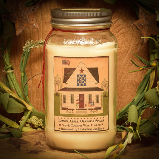 A 24 oz Mason jar candle with a label featuring a house and the scent of Lemon, Apple, Orange & Spices. The candle is set against a wooden background with greenery.