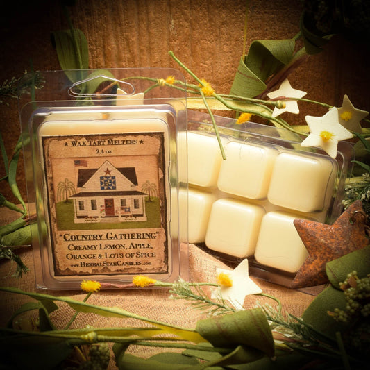 A package of Country Gathering mini wax tarts with a label depicting a rustic house scene. The tarts are visible in a clear plastic case, and there are decorative stars and foliage around the package.