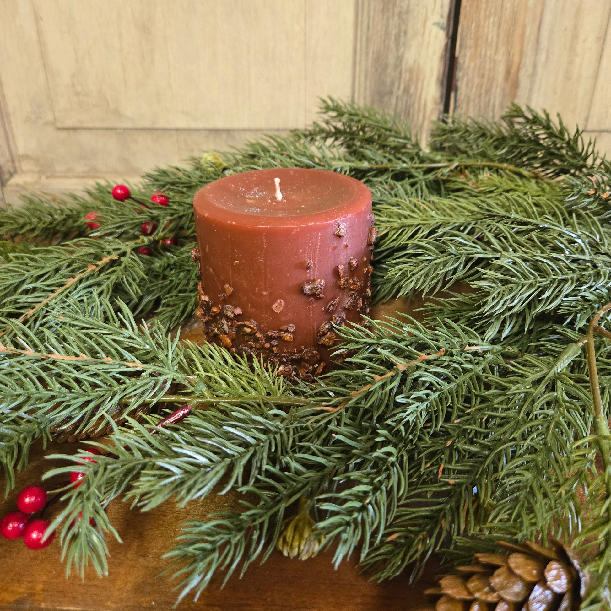 A pillar candle with a red label, surrounded by green pine branches and red berries on a wooden surface.